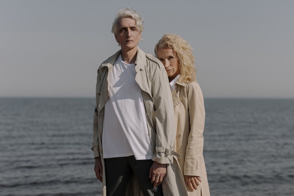 Man in White Dress Shirt Beside Woman in Brown Coat Standing on Seashore